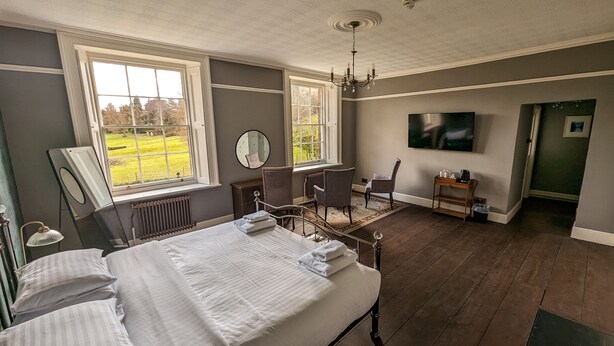 A bedroom with a large bed, two windows, a chandelier, a desk, and a TV. - Oakhill  (Matlock)