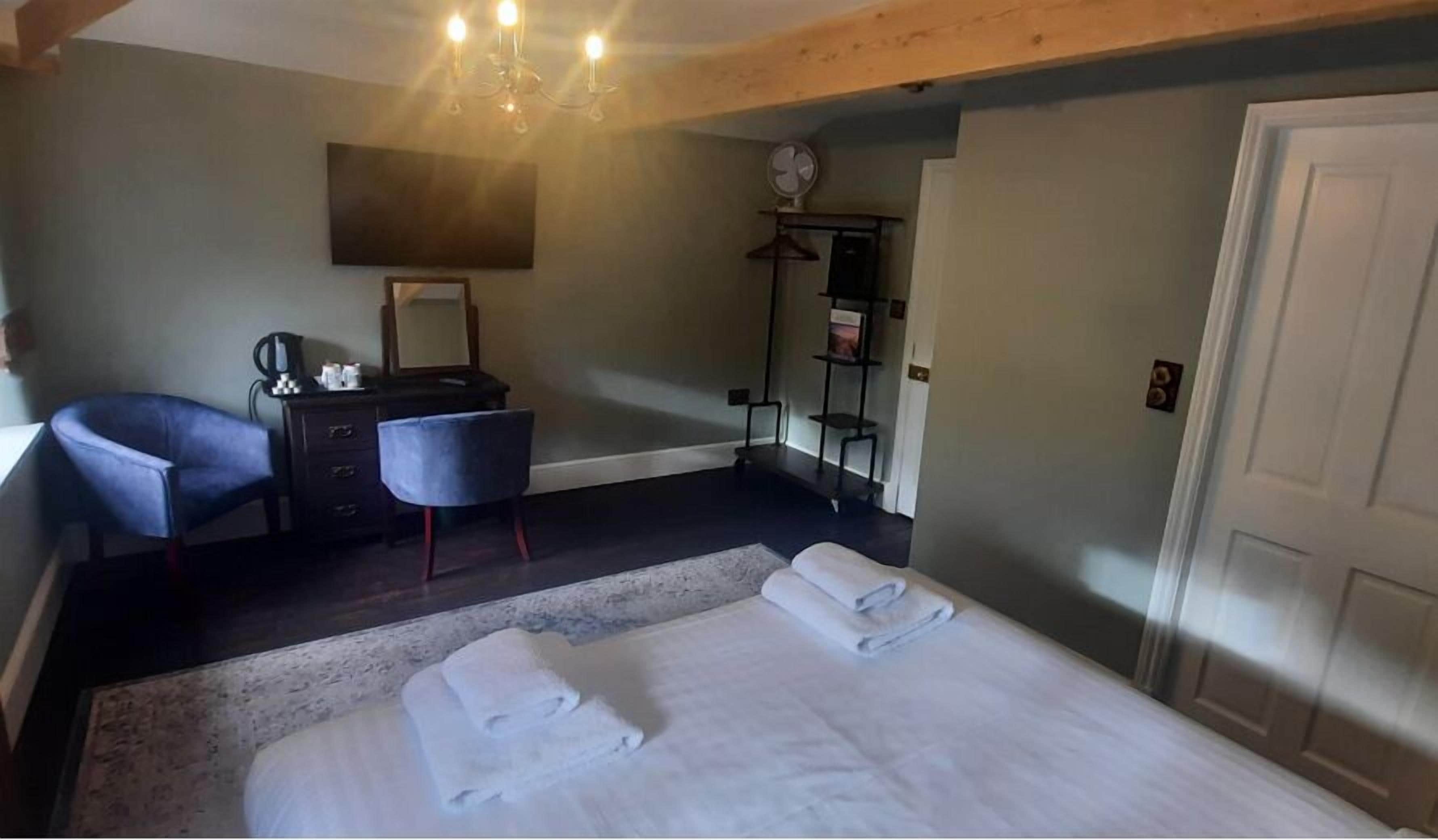 A bedroom with a bed, two chairs, a dresser, a mirror, a fan, and a shelf with items.