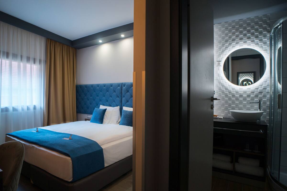 A hotel room with a large bed, a bathroom with a modern sink and mirror, and a view of the city through the window.