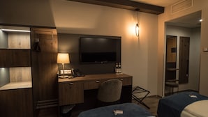 A hotel room with a wooden desk, a flat-screen TV, a desk lamp, two beds, and a wardrobe.