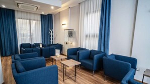 A modern hotel room with blue upholstered sofas arranged around a central coffee table, large windows with sheer curtains, and a visible fireplace.