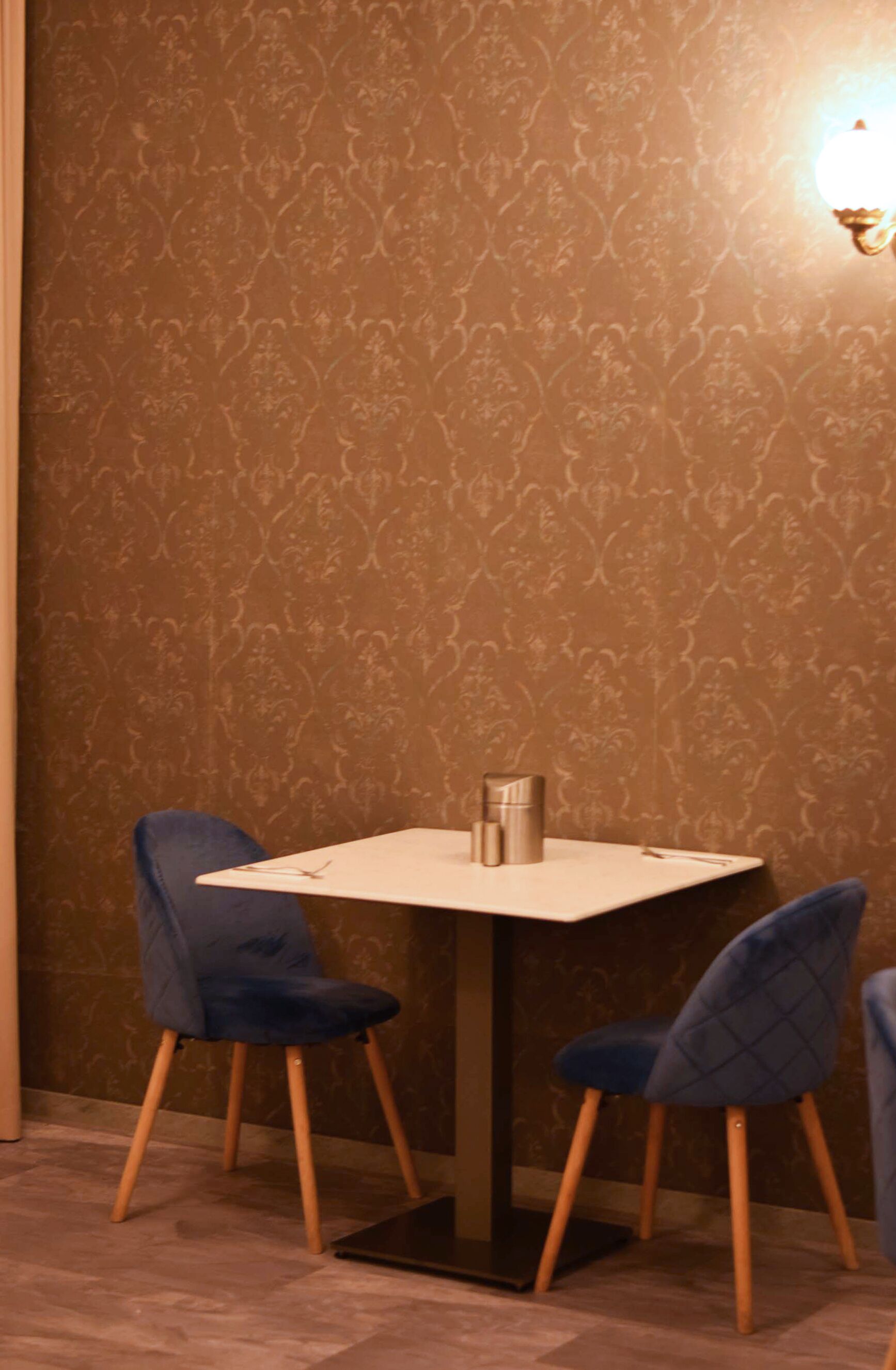 Uma área de refeições com uma mesa e duas cadeiras, um objeto cilíndrico pequeno sobre a mesa e uma parede com um design estampado.