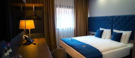 A hotel room with a bed, a desk with a telephone, a chair, and a window with curtains.