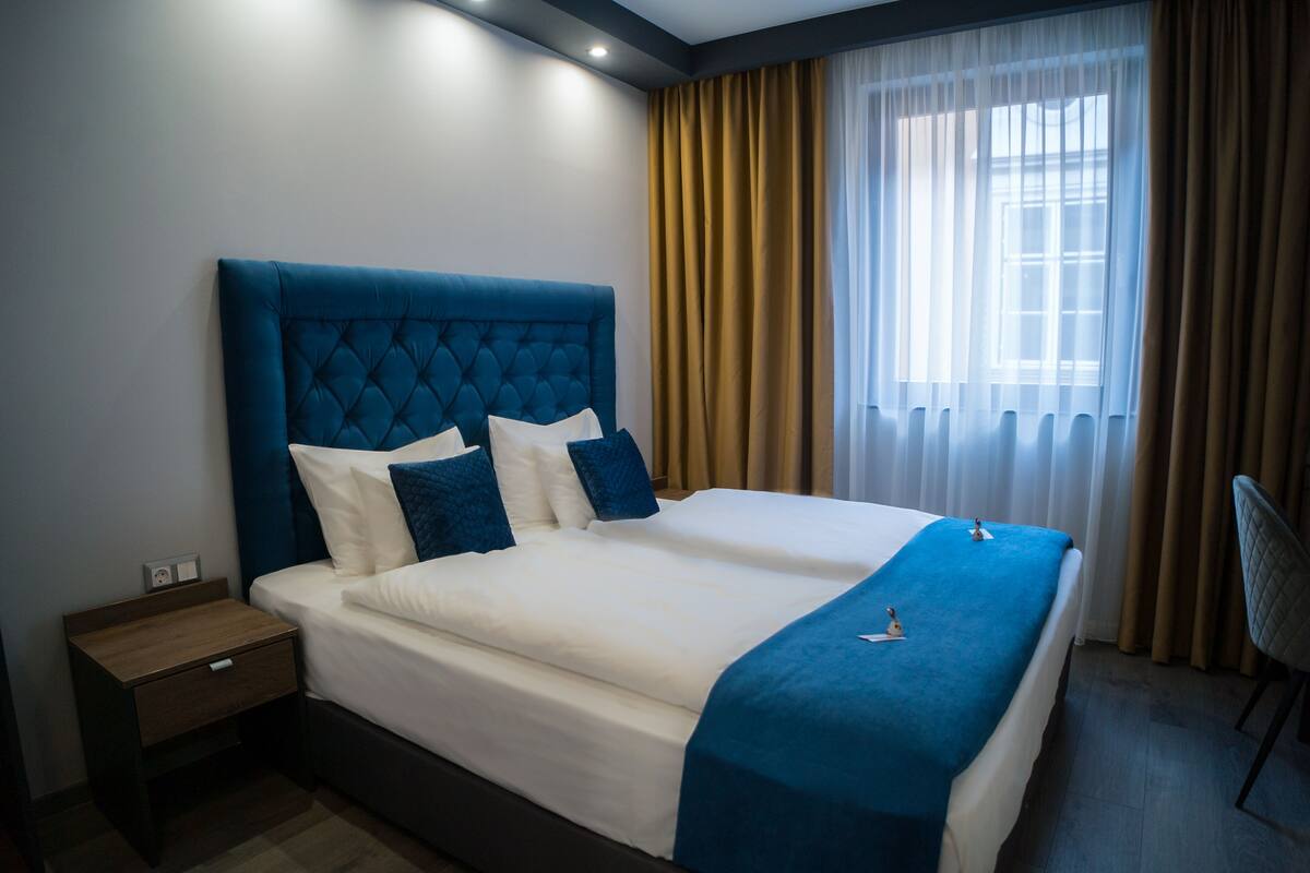 A double bed with a tufted headboard, two pillows, a bedside table, and a window with curtains.