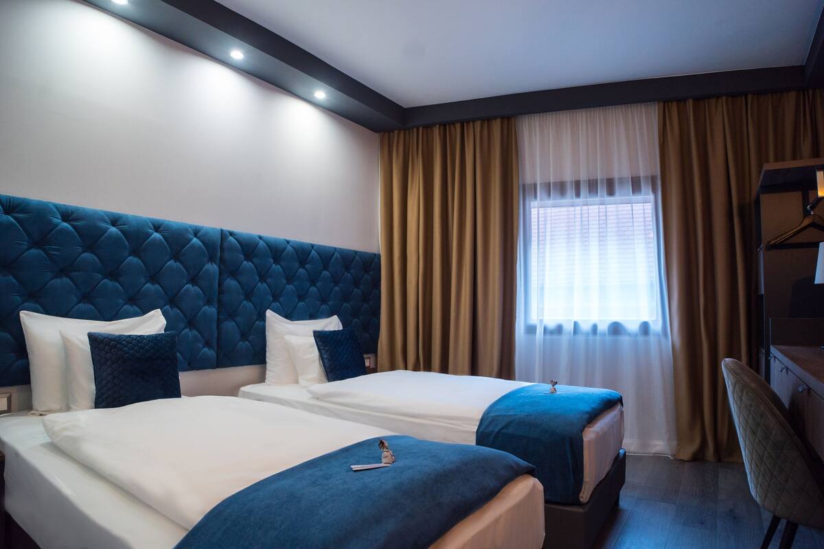 A hotel room with two beds, a large tufted headboard, a desk, and a window with curtains.