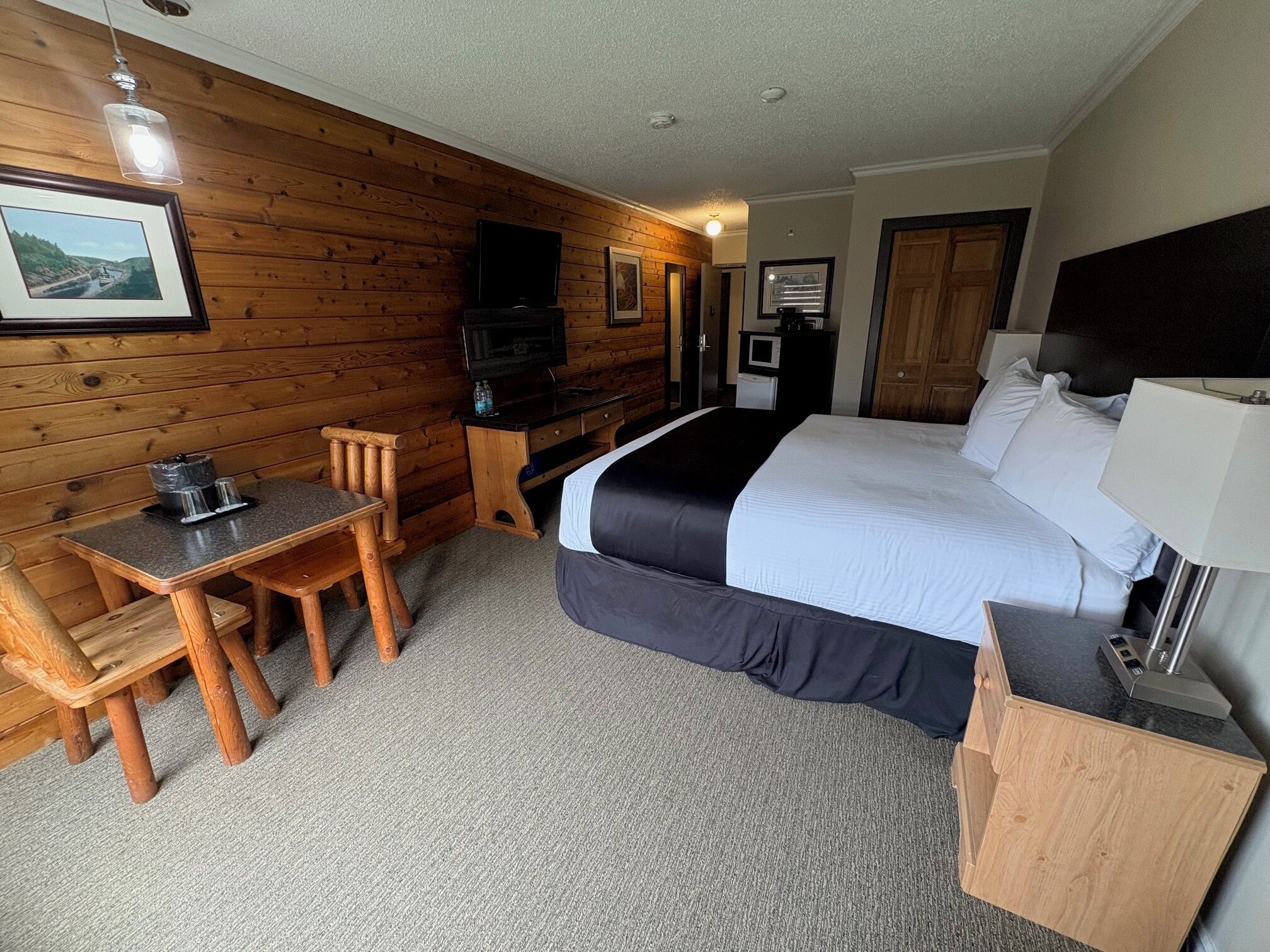 A hotel room with a wooden wall, a bed, a desk, a chair, a TV, and a small kitchenette.