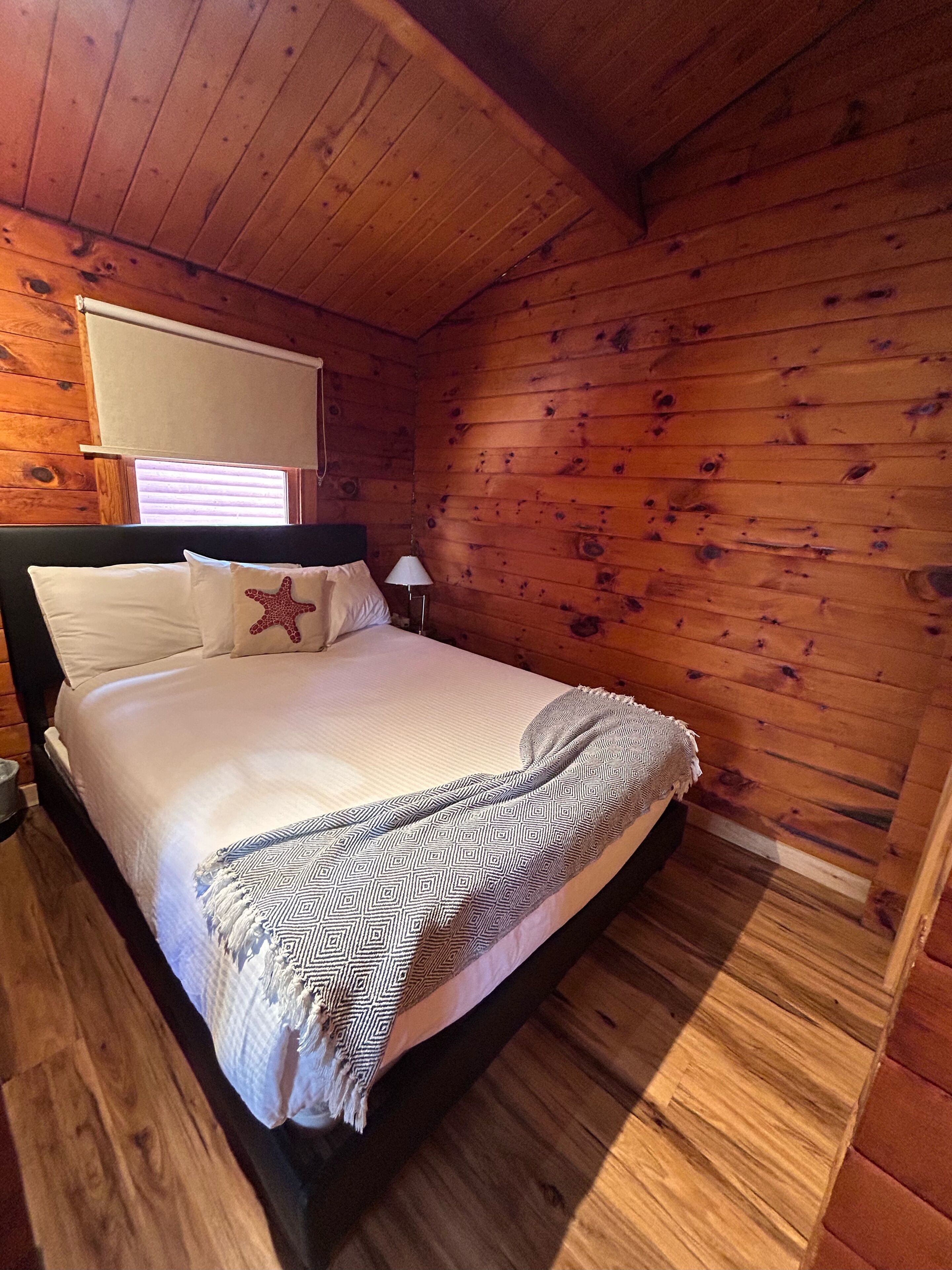 A wooden cabin bedroom with a bed, a starfish pillow, a lamp, and a window with blinds.