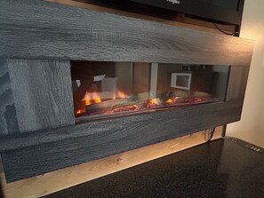 A fireplace with a dark wood frame and a glass front, displaying a fire inside. - Bras d'Or Lakes Inn (St. Peters)