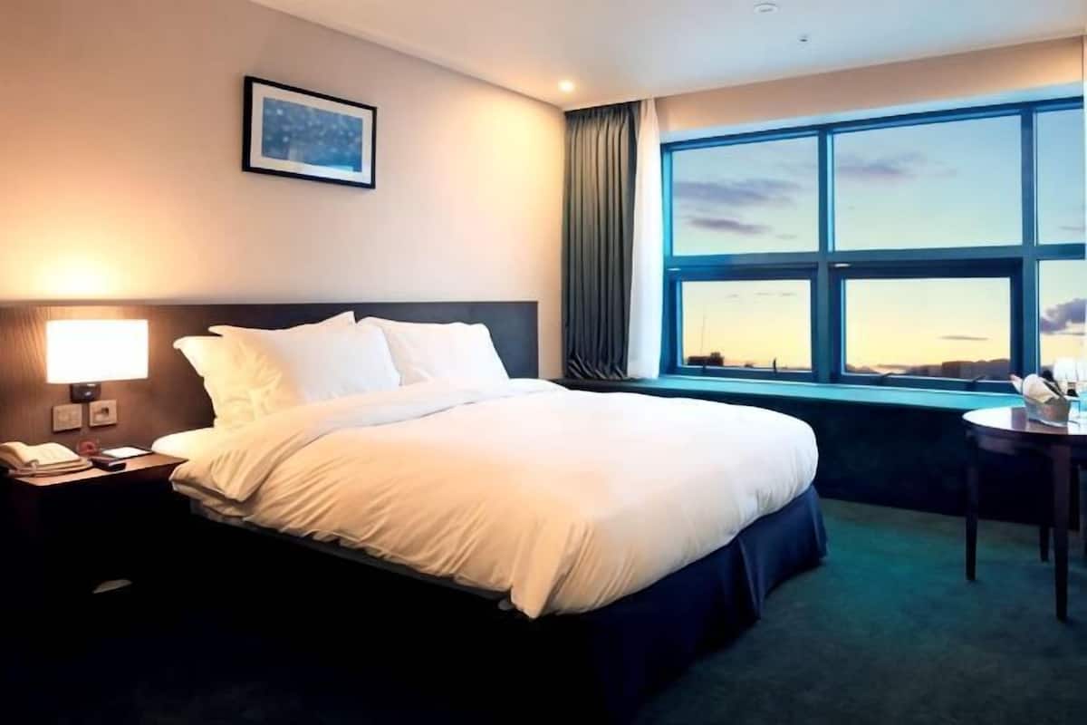 Ett hotellrum med en stor säng, ett nattduksbord med en lampa, ett skrivbord med en stol och ett fönster med utsikt.