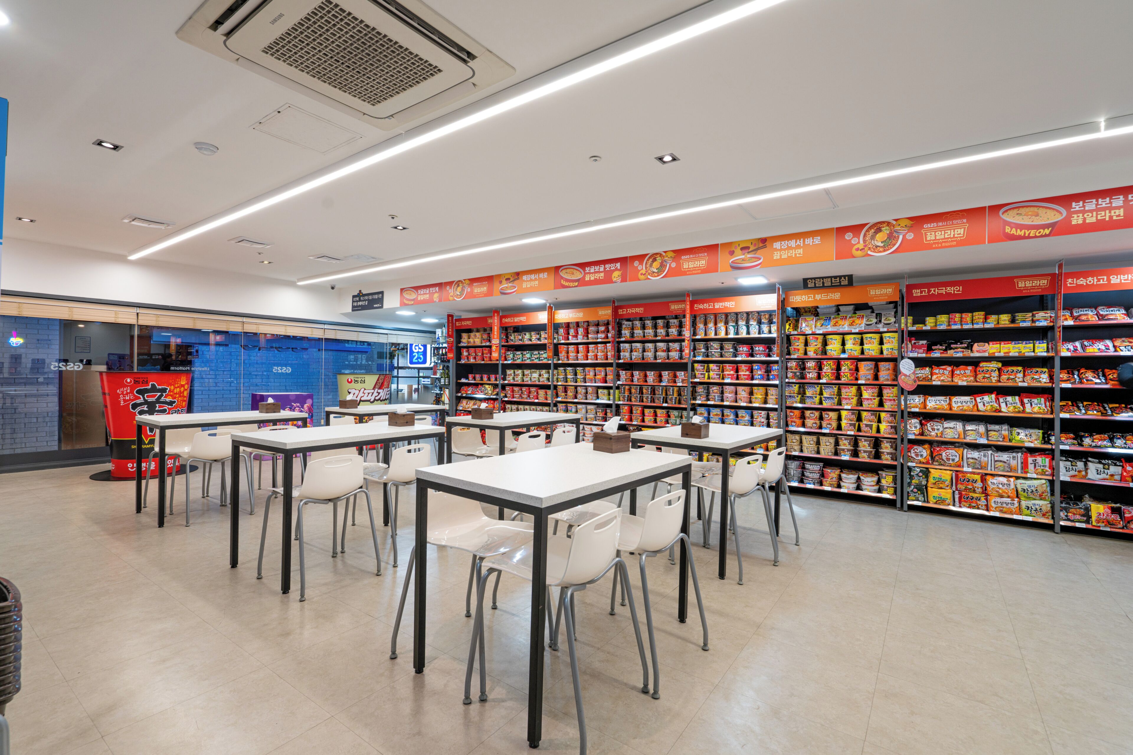 A cafeteria with tables and chairs, shelves stocked with food items, and a blue wall with a sign.