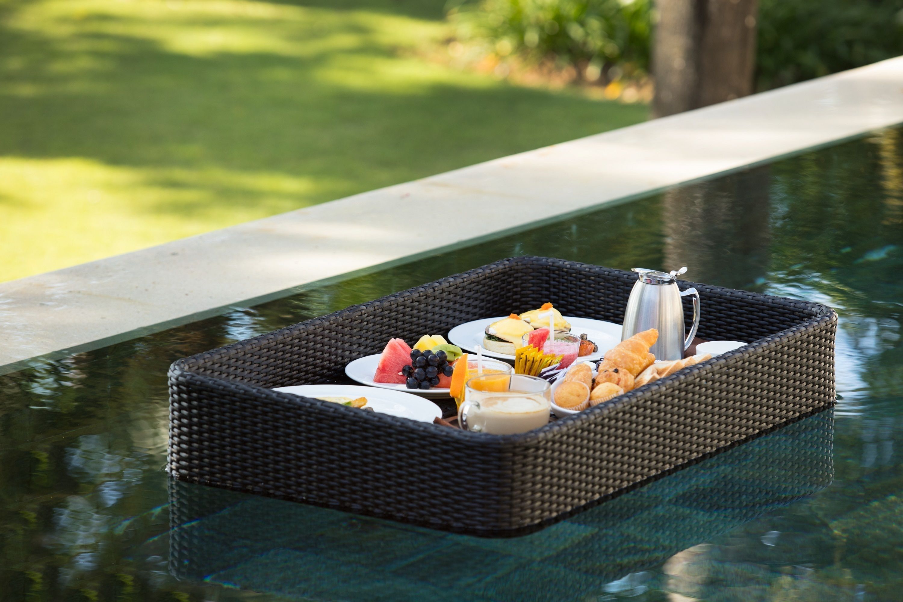 A wicker tray with breakfast items on a glass table near a pool.