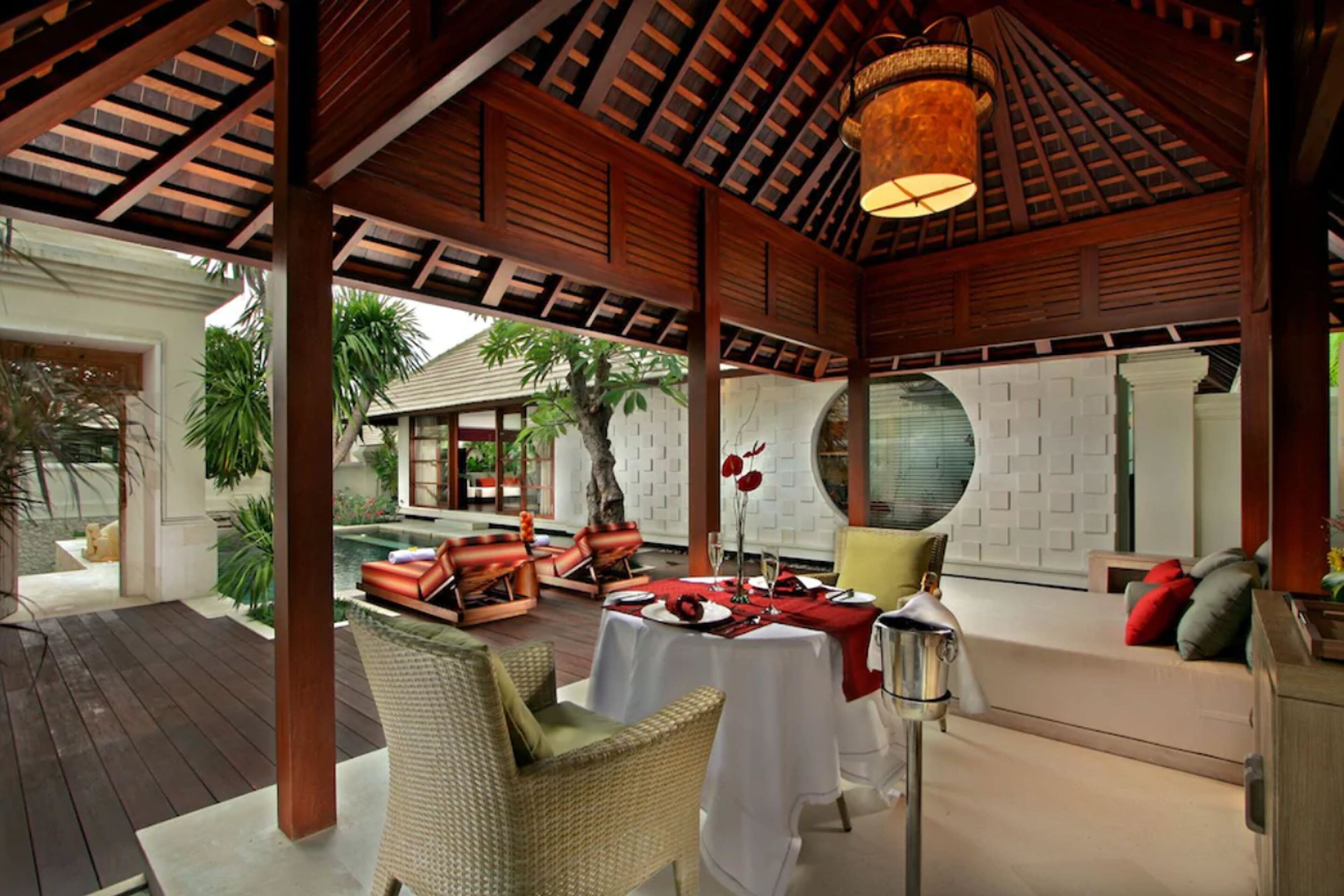 A covered outdoor dining area with wooden beams, a pool, and a view of a white building with a tiled roof.