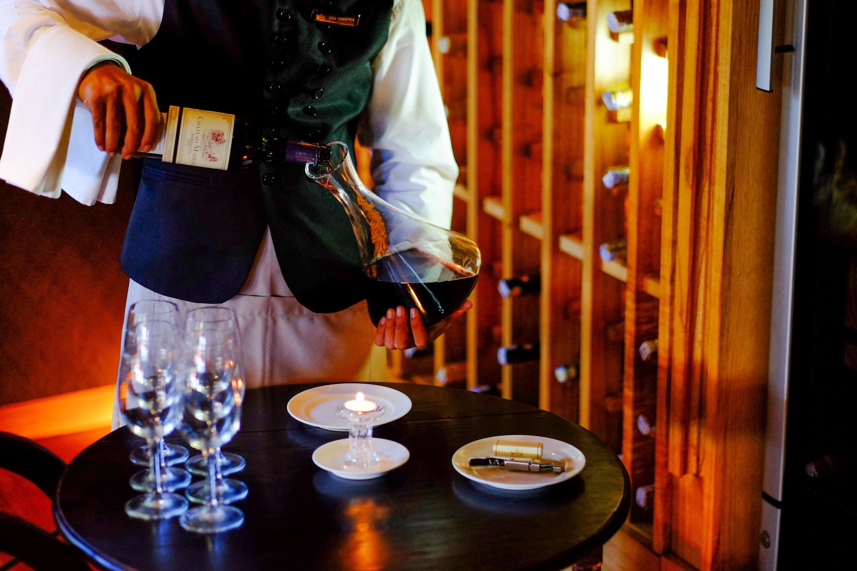 A person pouring wine into glasses on a table with a candle and a plate of cheese.