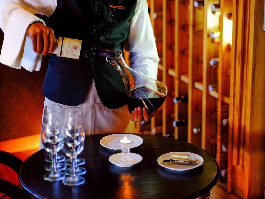 A person pouring wine into glasses on a table with a candle and a plate of cheese.