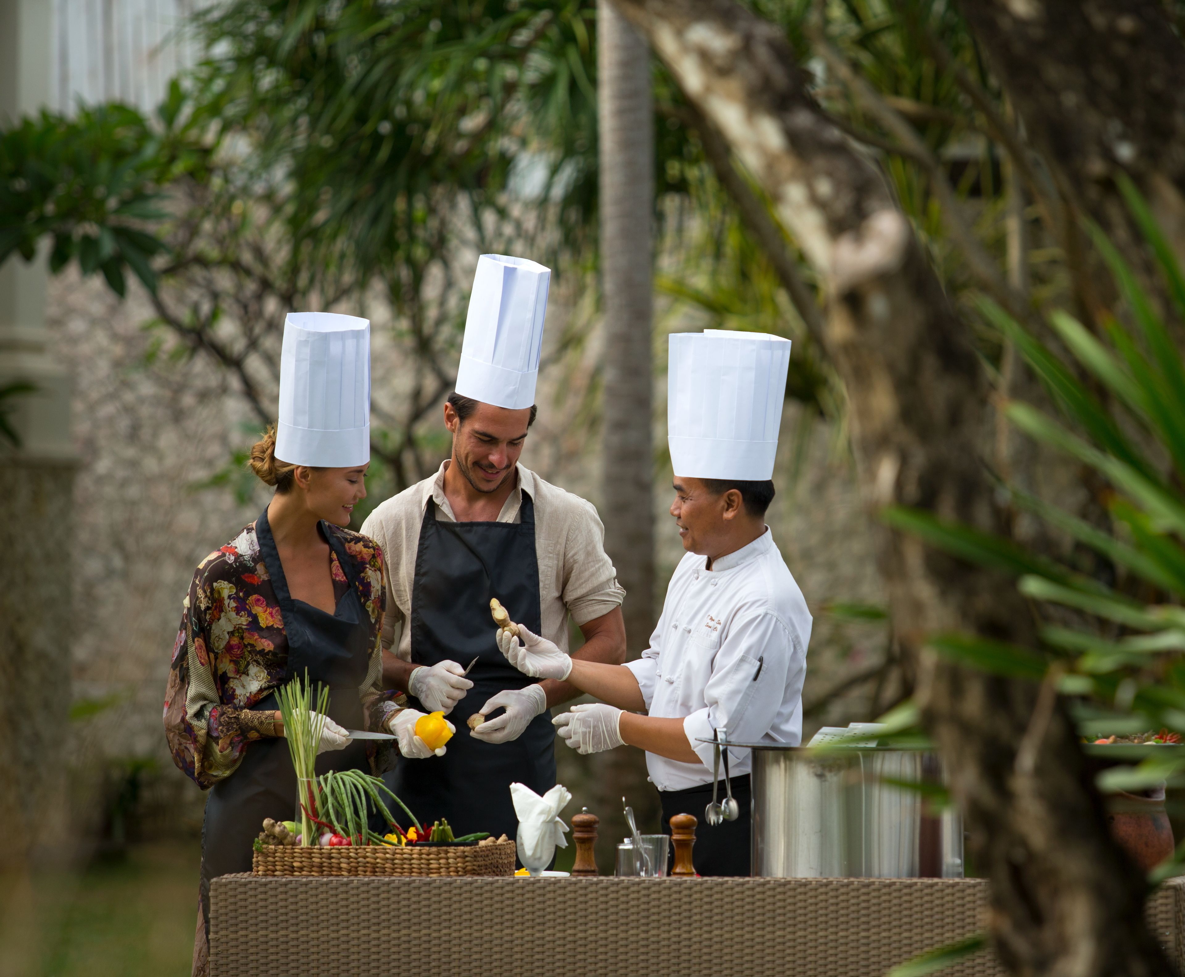 Three people wearing chef hats and aprons, preparing food outdoors with a basket of vegetables and cooking utensils on a table.