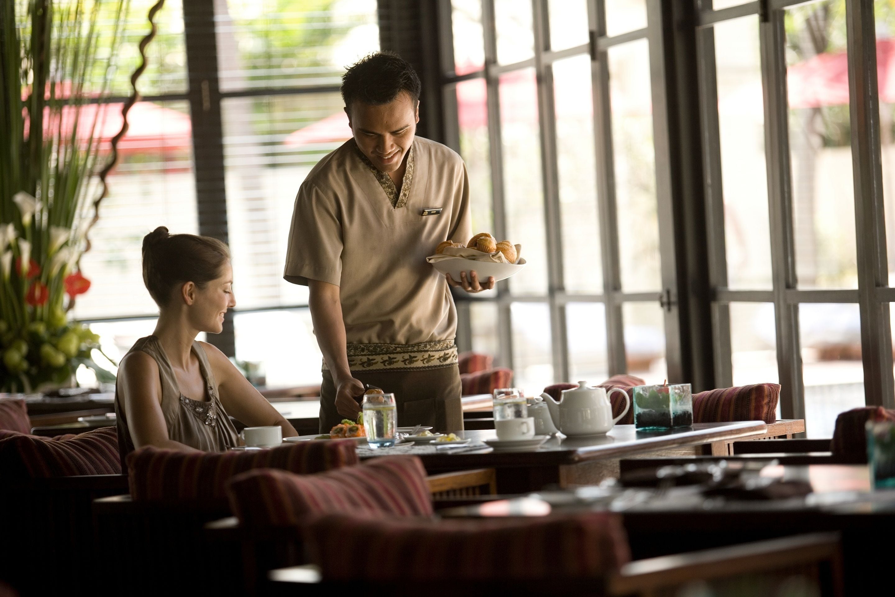 A person serving food to a seated individual in a well-lit room with large windows, a table set with dishes, and a relaxed seating area.
