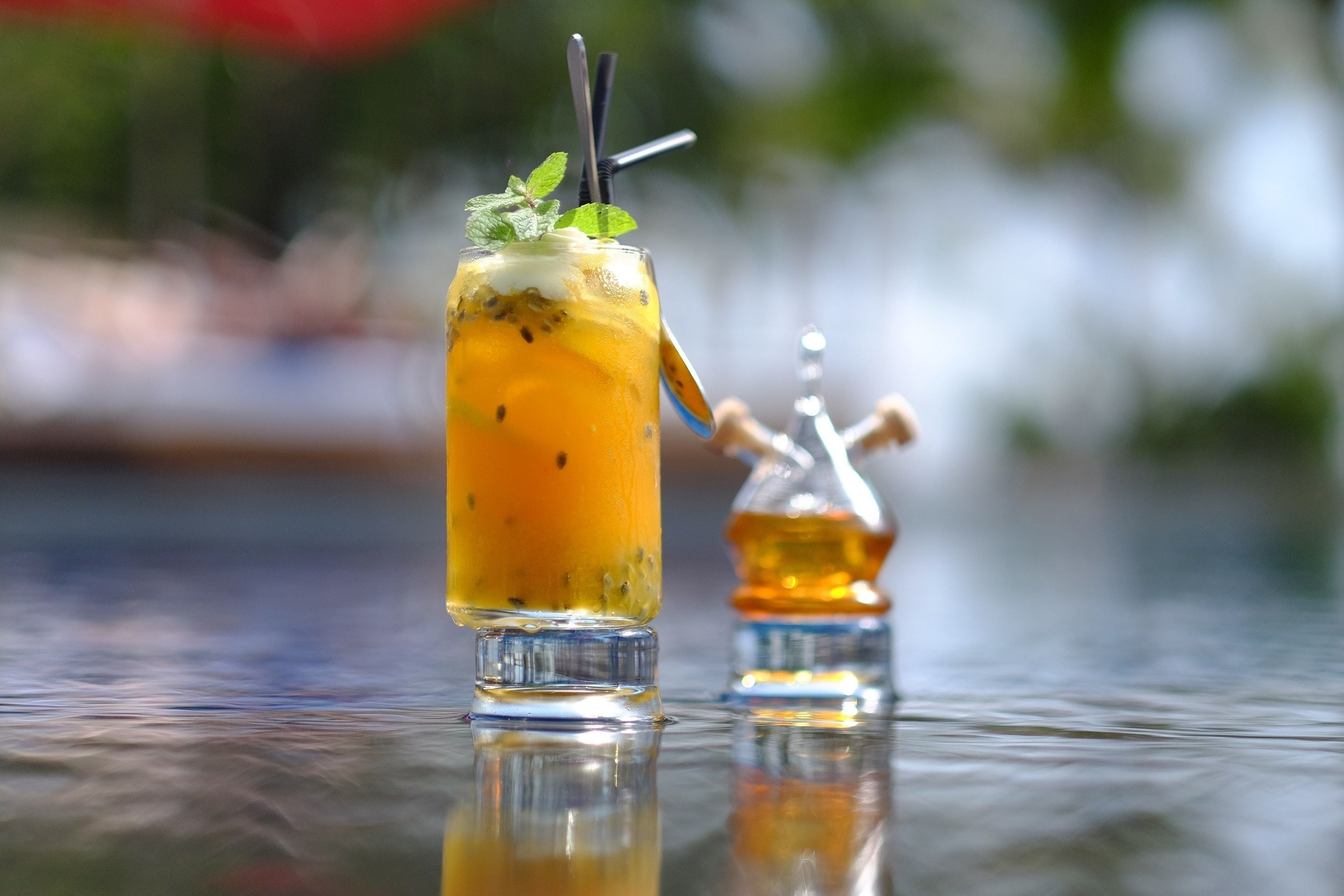 A glass of yellow beverage with mint garnish and a straw, placed on a reflective surface with a blurred background.