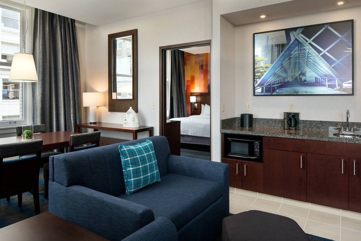A hotel room with a sofa, a dining table, a kitchen area, and a large window.