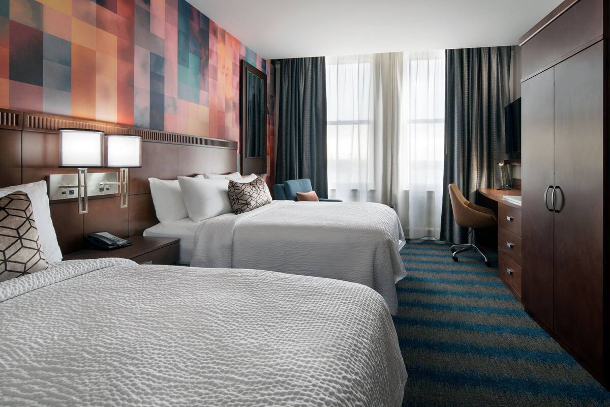 A hotel room with two beds, a desk, a chair, a TV, and a large window with curtains.