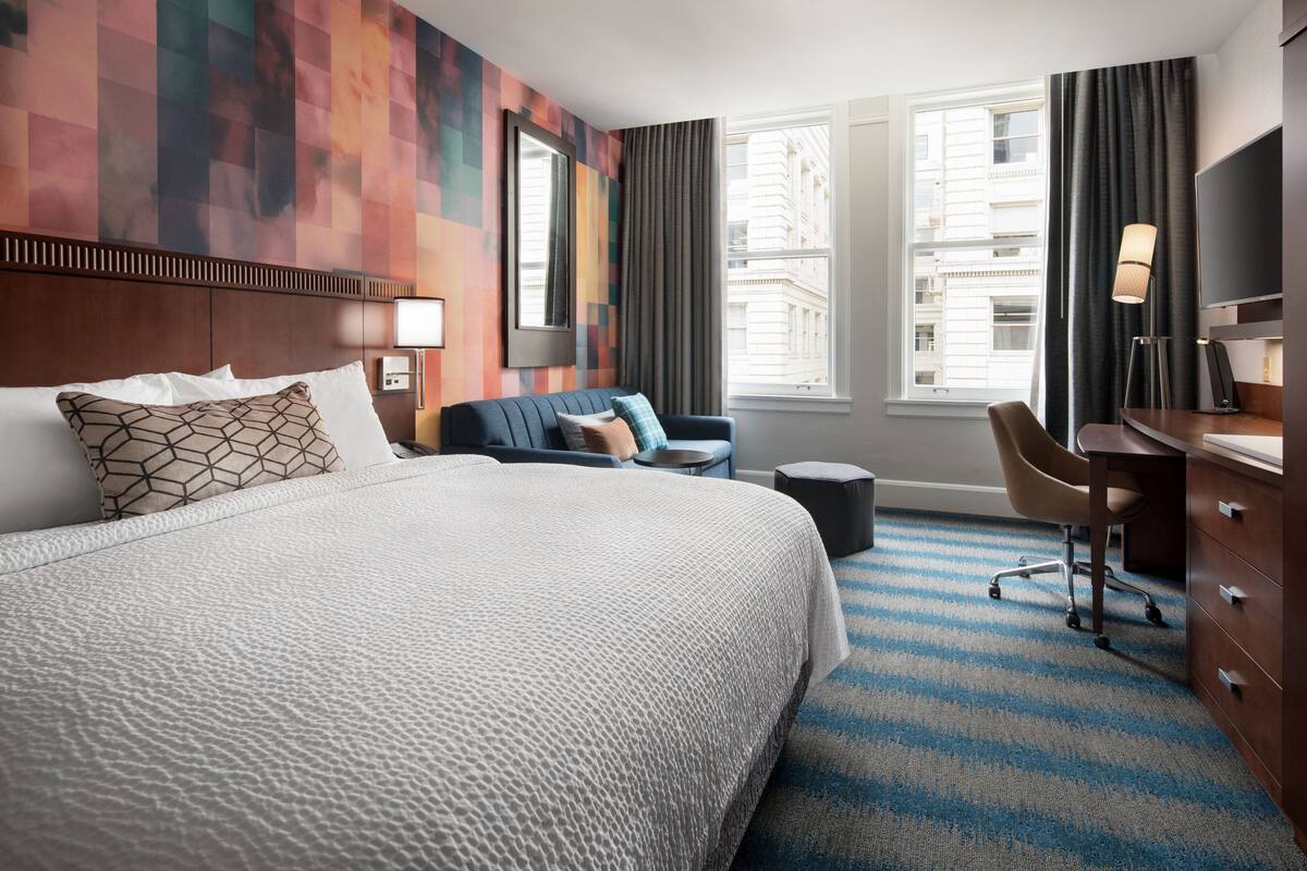 A hotel room with a large bed, a desk, a chair, and a view of the city through the window.