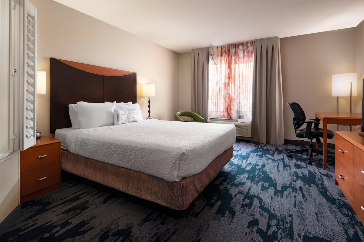 A hotel room with a large bed, a desk, a chair, a lamp, and a window with curtains.