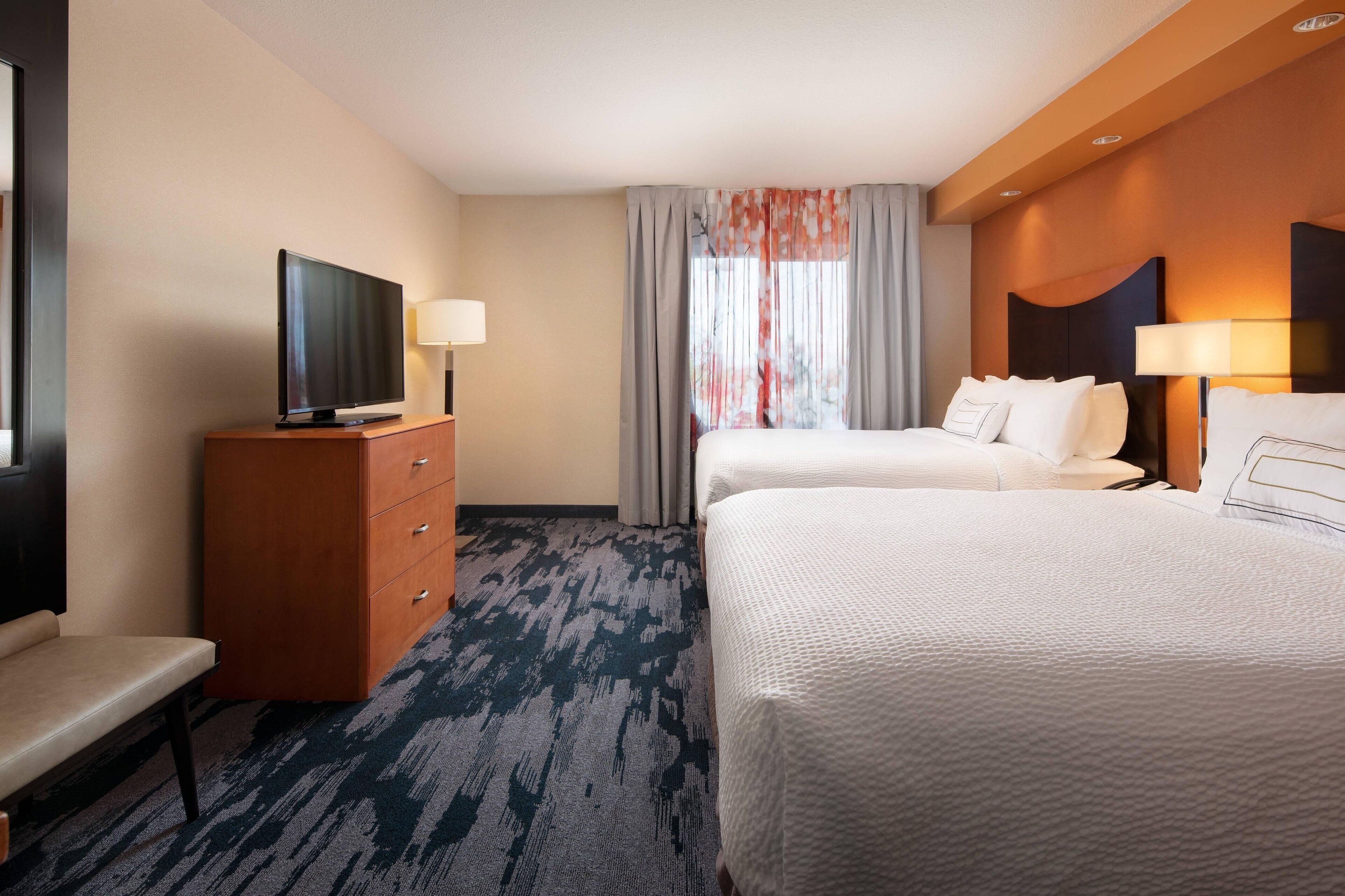 Une chambre d’hôtel avec un téléviseur à écran plat posé sur une commode en bois, deux lits avec du linge de lit blanc, un bureau avec une lampe et une grande fenêtre ornée de rideaux légers.