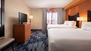 Une chambre d’hôtel avec un téléviseur à écran plat posé sur une commode en bois, deux lits avec du linge de lit blanc, un bureau avec une lampe et une grande fenêtre ornée de rideaux légers.