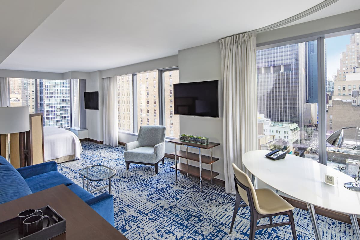 A hotel room with a large window, a flat-screen TV, a dining table, and a sofa.