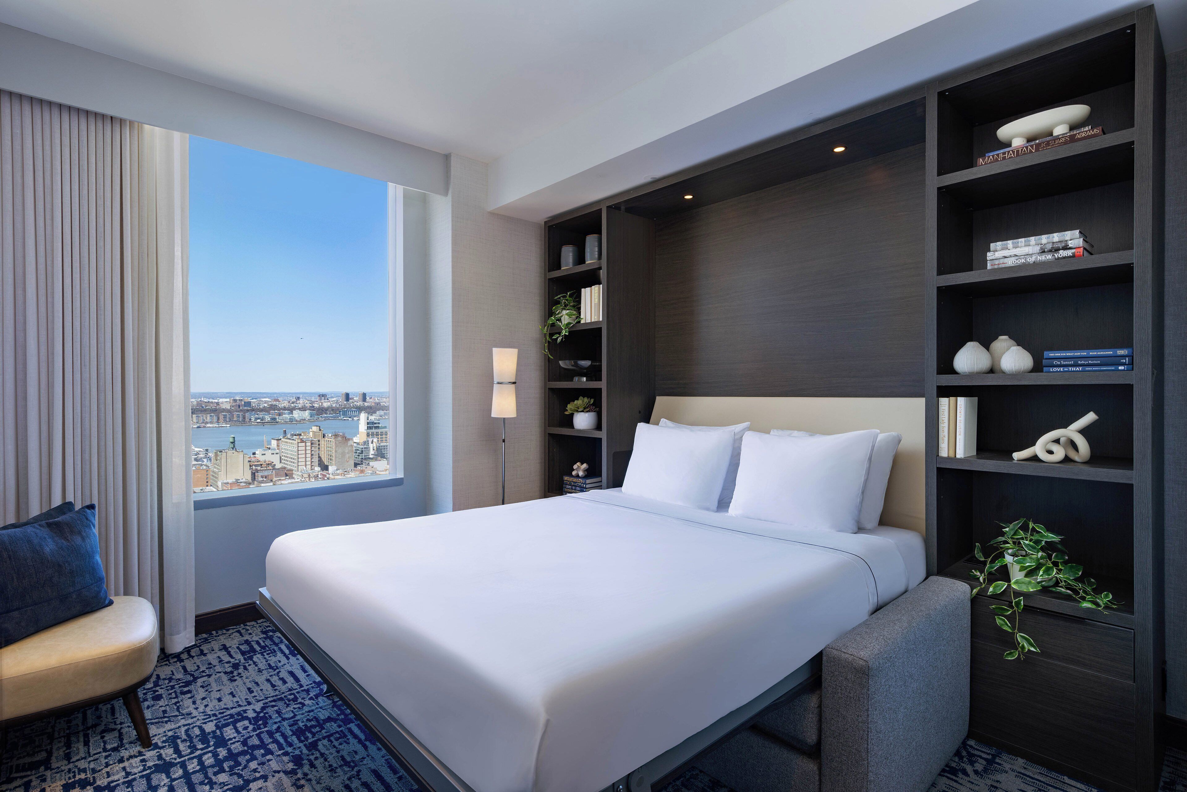 A hotel room with a large bed, a view of the city, a bookshelf, and a desk area.
