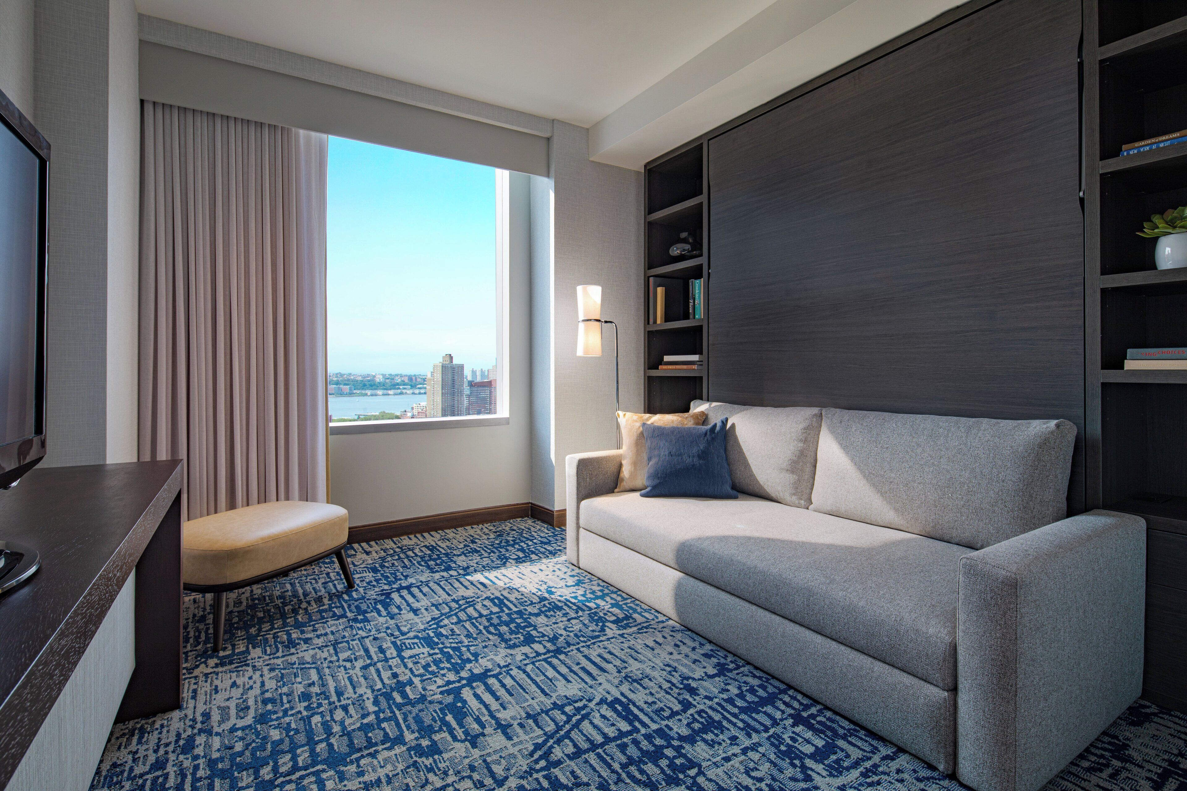 A modern hotel room with a sofa, a television, a bookshelf, and a large window offering a city view.