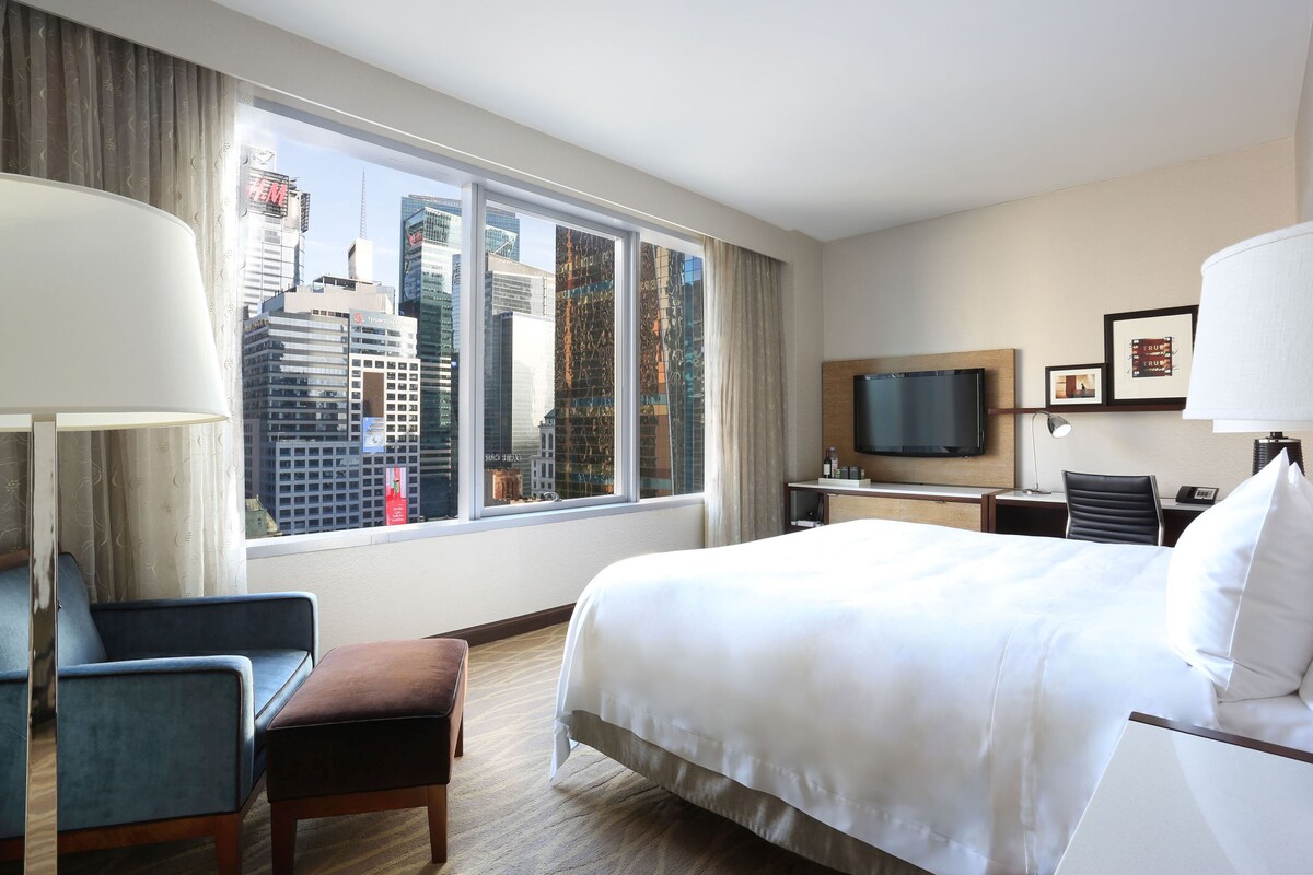 A hotel room with a large bed, a desk, a chair, a TV, and a window with a city view.