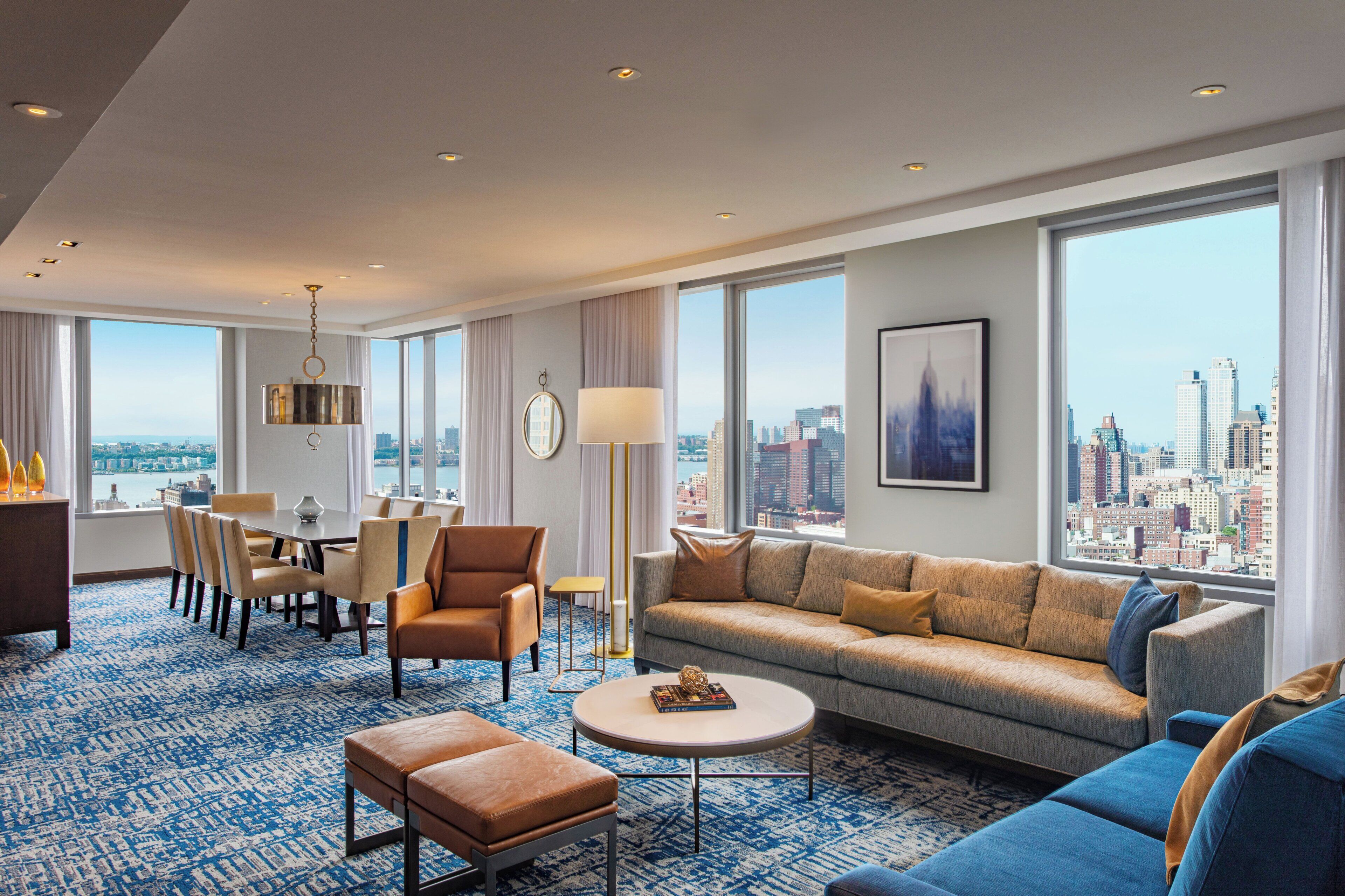 A modern hotel room with a large window offering a city view, a dining area with a table and chairs, a sofa, a coffee table, and a floor lamp.