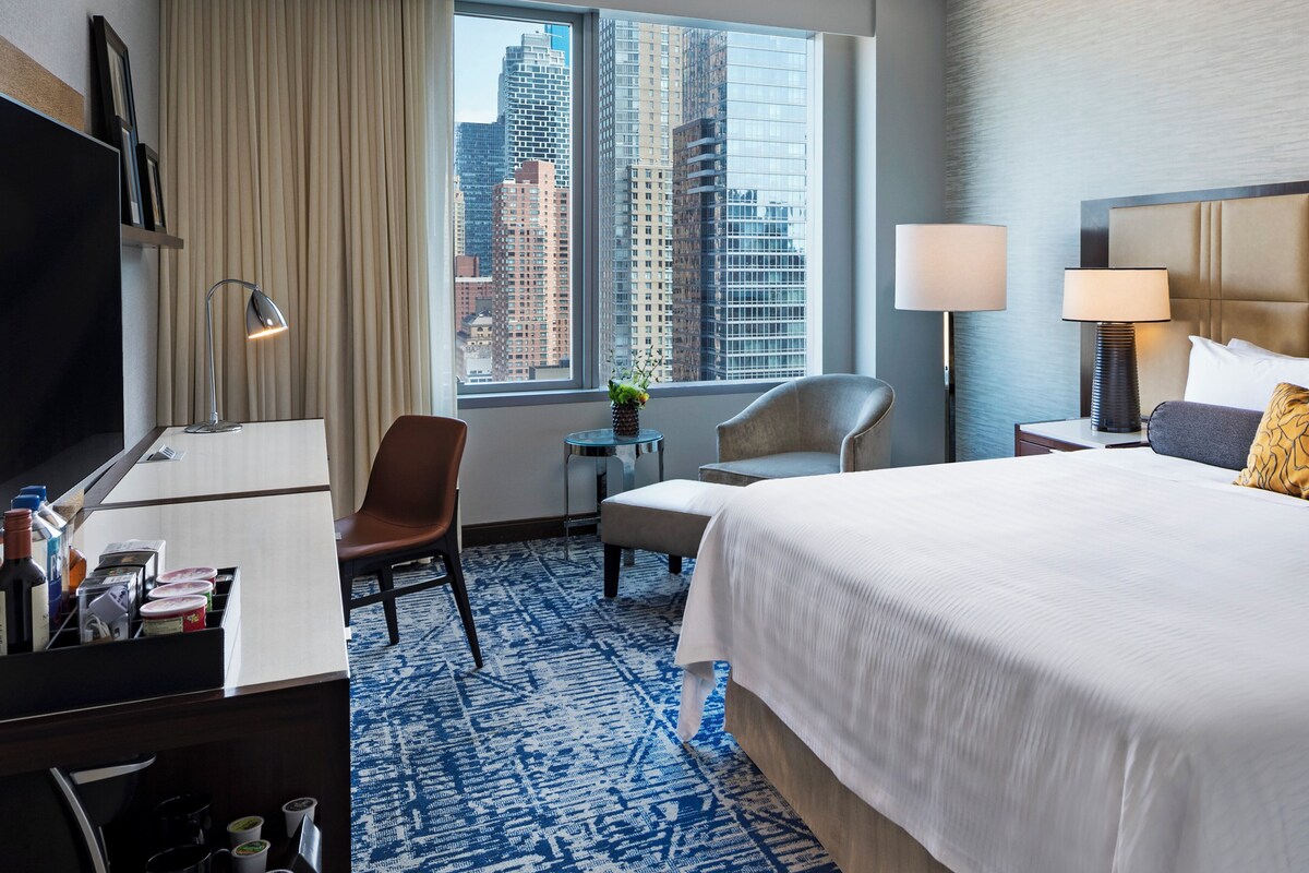 A hotel room with a large bed, a desk with a lamp, a chair, a small table with a plant, and a view of tall buildings through the window.
