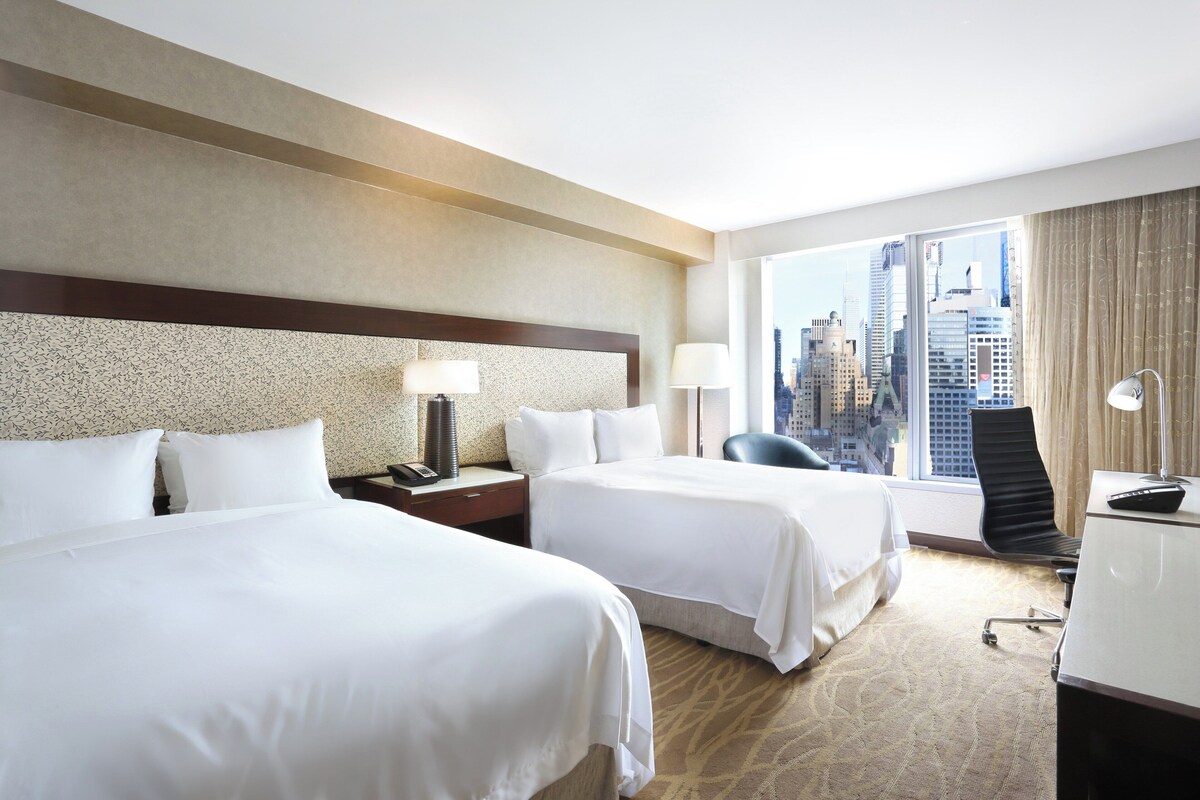 A hotel room with two beds, a desk, a chair, and a large window offering a city view.