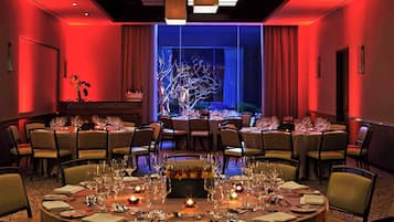 A hotel dining area with round tables set for a meal, a large window with a view of a tree, and red ambient lighting.