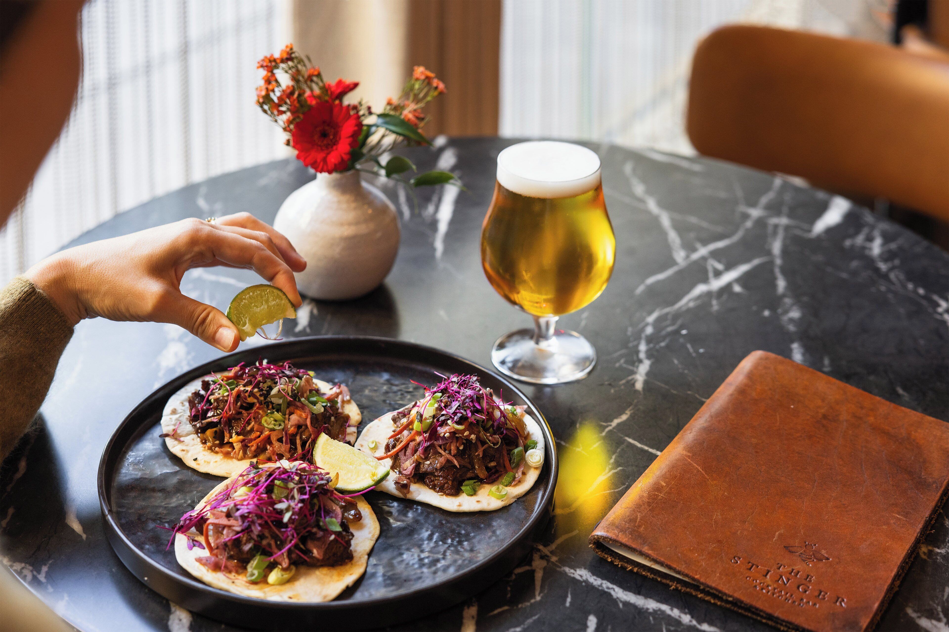 Uma mesa com três tacos, um copo de cerveja e um vaso de flores.