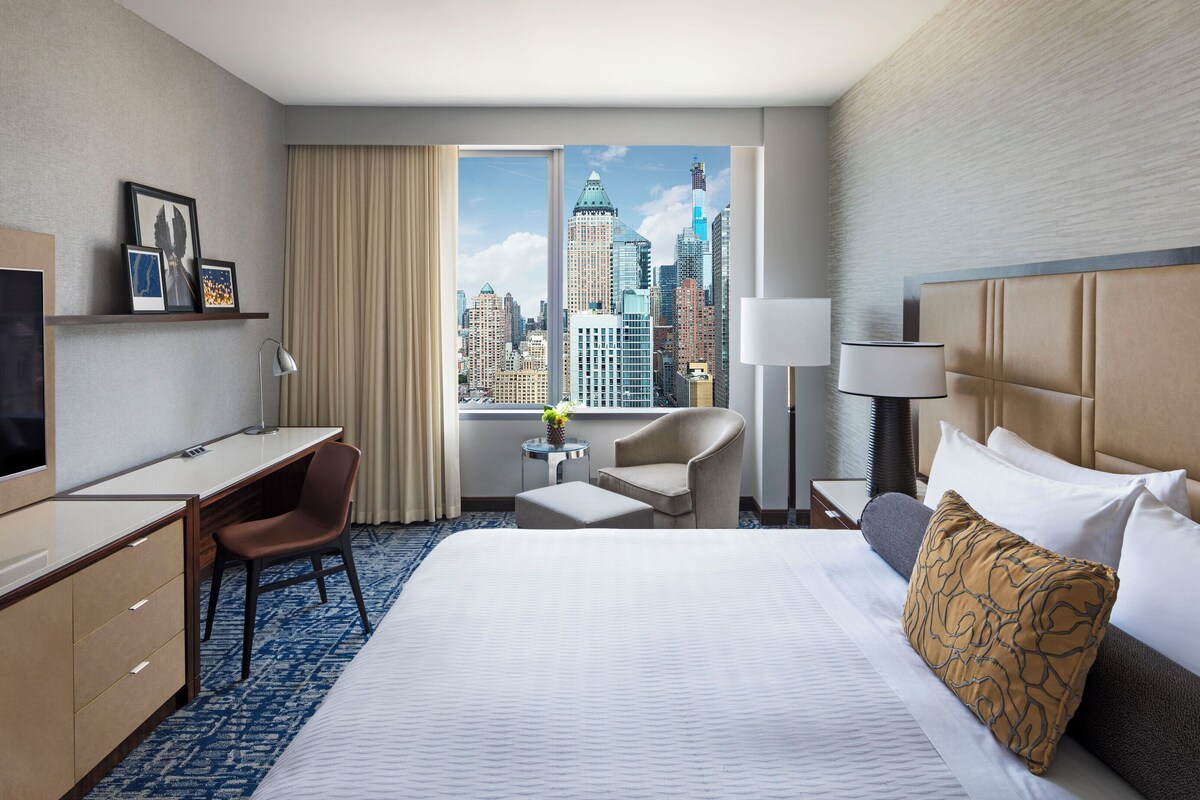 A hotel room with a bed, desk, chair, TV, and a view of a cityscape through large windows.