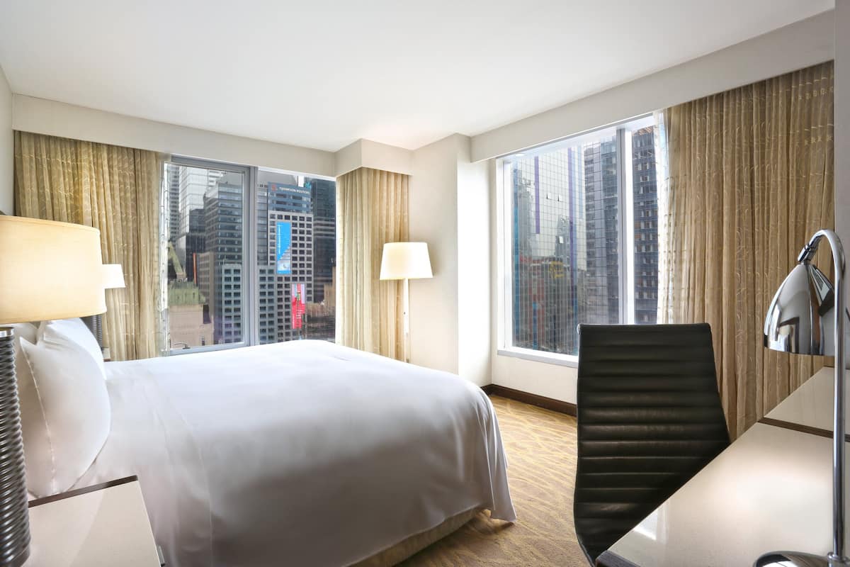 A hotel room with a large bed, a desk, a chair, two lamps, and large windows offering a city view.