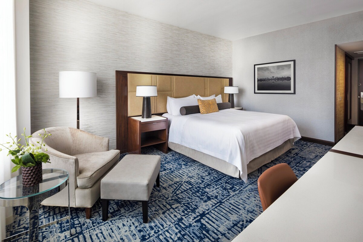 A hotel room with a bed, two bedside lamps, a nightstand, a chair, a small table with a vase, and a blue patterned carpet.