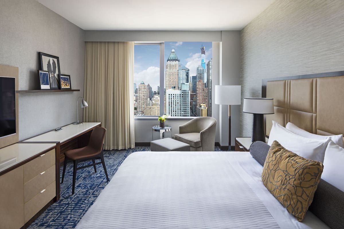 A hotel room with a bed, desk, chair, TV, and a view of a cityscape through large windows.