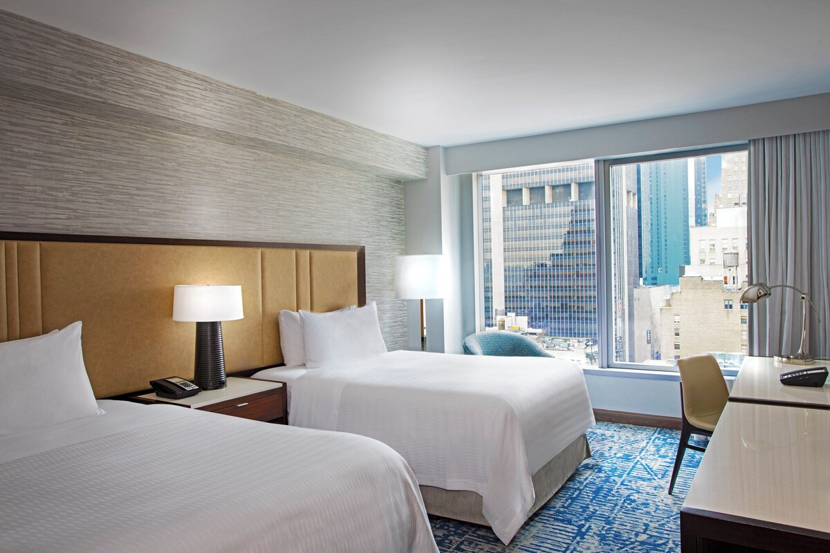 A hotel room with two beds, a desk, a chair, and a large window offering a city view.