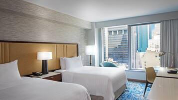 A hotel room with two beds, a desk, a chair, and a large window offering a city view.