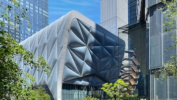 Un edificio moderno con una fachada geométrica distintiva, rodeado de vegetación y otros edificios de gran altura.