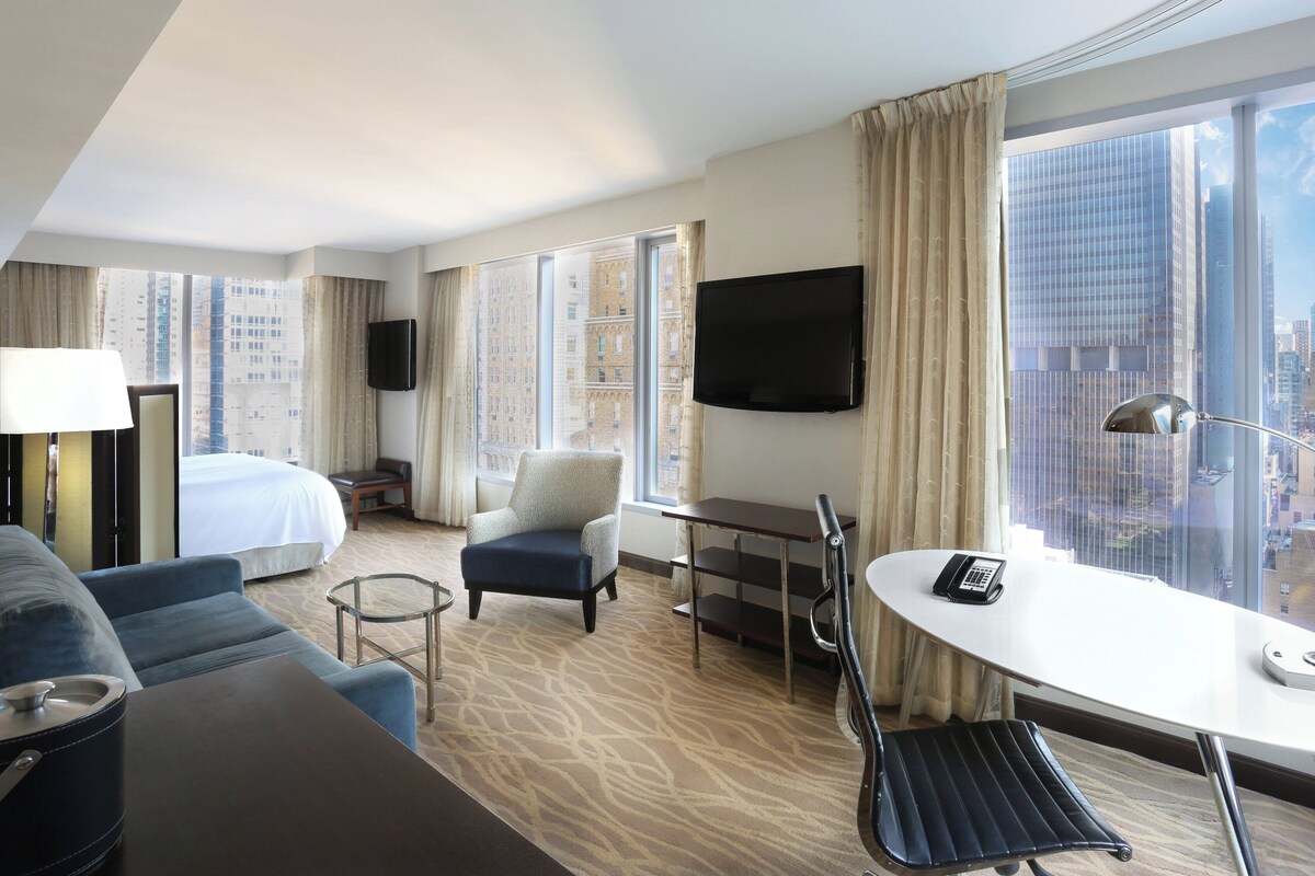 A hotel room with a bed, a sofa, a desk with a telephone, a chair, a TV, and a large window with a city view.