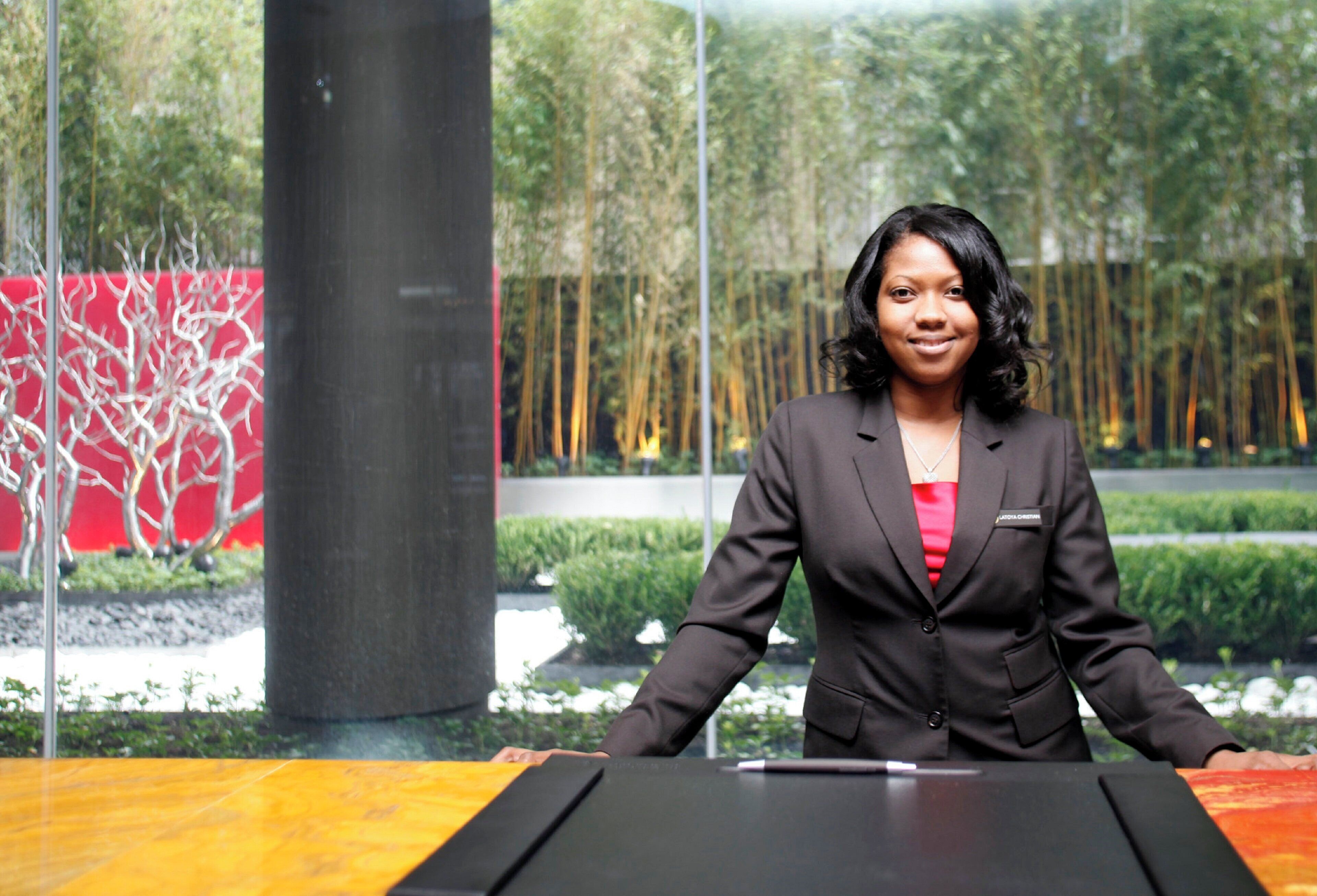 A person standing at a podium with a name tag, in a modern indoor setting with glass walls and a view of greenery outside.