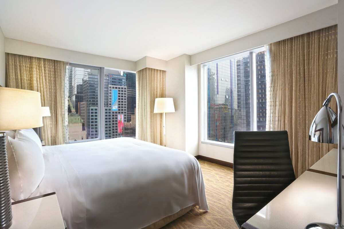 A hotel room with a large bed, a desk, a chair, two lamps, and large windows offering a city view.