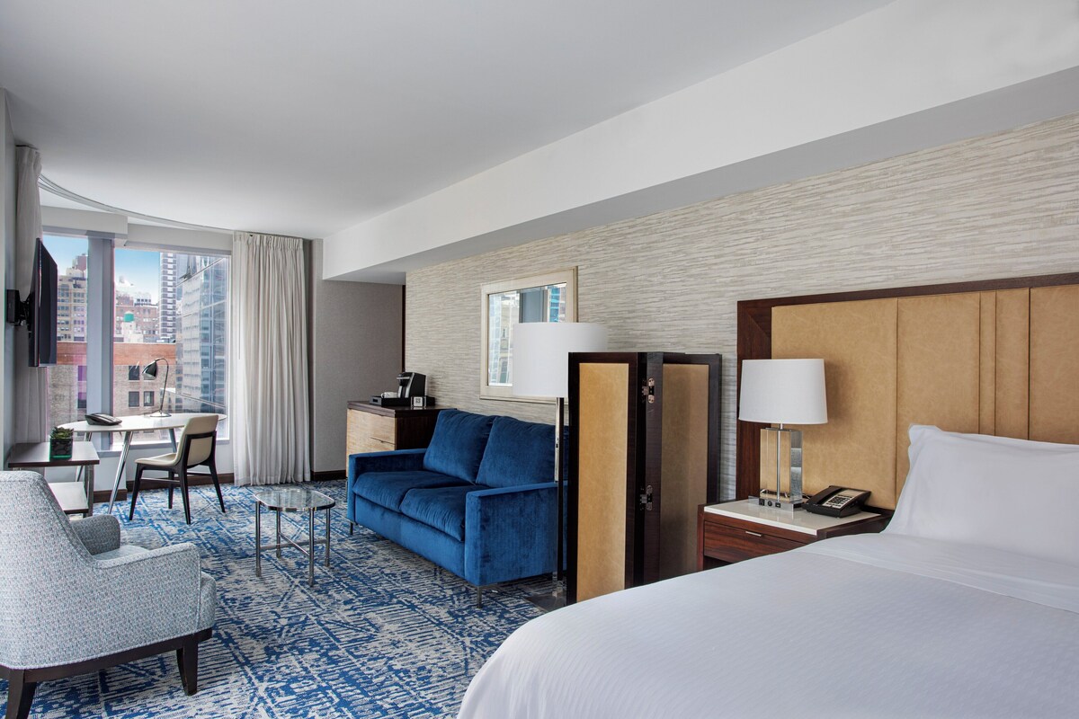 A hotel room with a bed, a desk, a sofa, a chair, and a view of the cityscape.