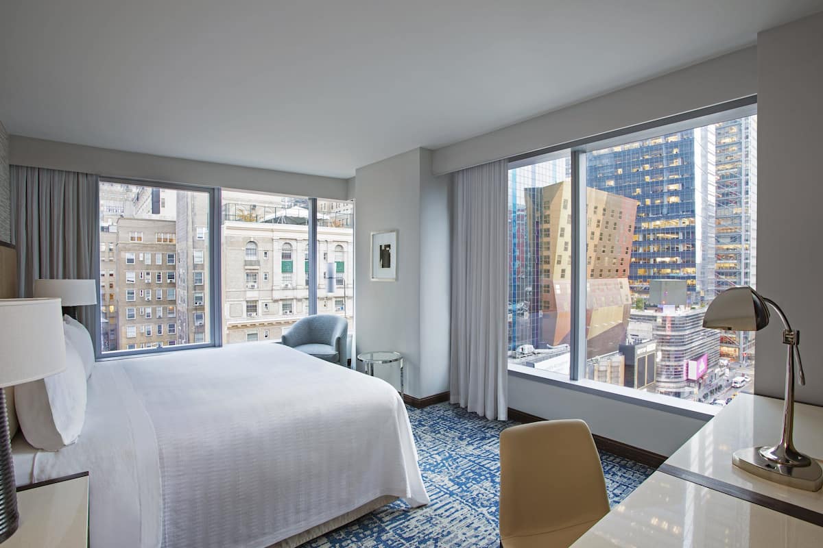 A hotel room with a large bed, a desk, a chair, and a view of the cityscape through large windows.