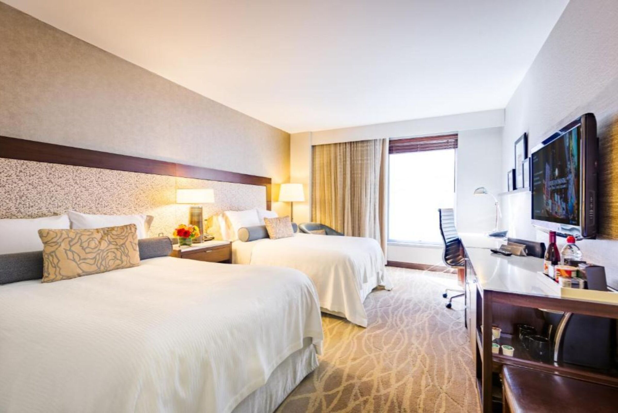 A hotel room with two beds, a desk, a TV, and a large window.