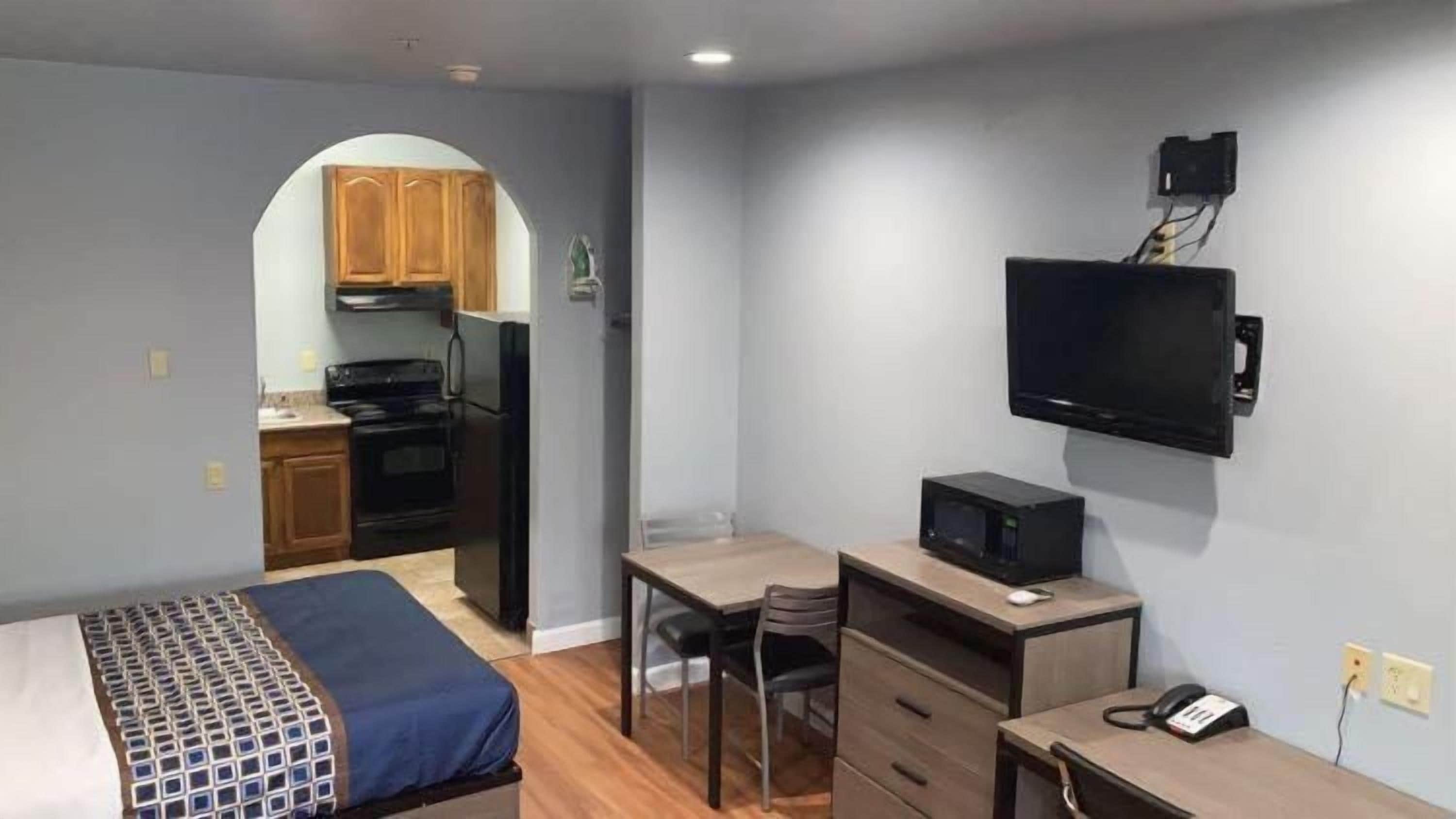 Quarto compacto com cama, escrivaninha, cadeira, televisão e cozinha compacta.